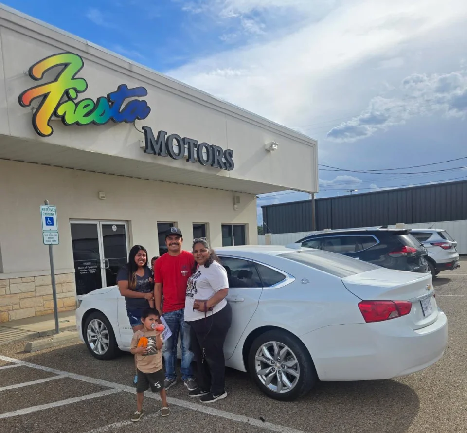 A happy family stands next to their newly purchased car in front of Fiesta Motors, a buy here pay here used car dealership.